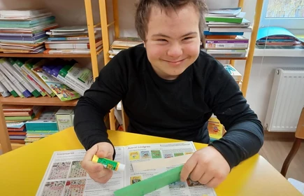 Chłopiec siedzi przy stoliku w bibliotece szkolnej, uśmiecha się, w te widać regały z książkami.