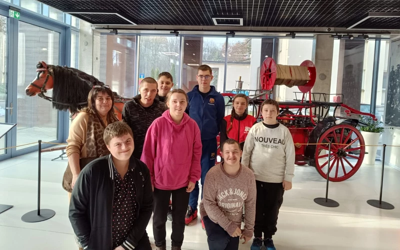 Grupa uczniów stoi w sali wystawowej muzealnej, w tle widać stary pojazd pożarniczy.