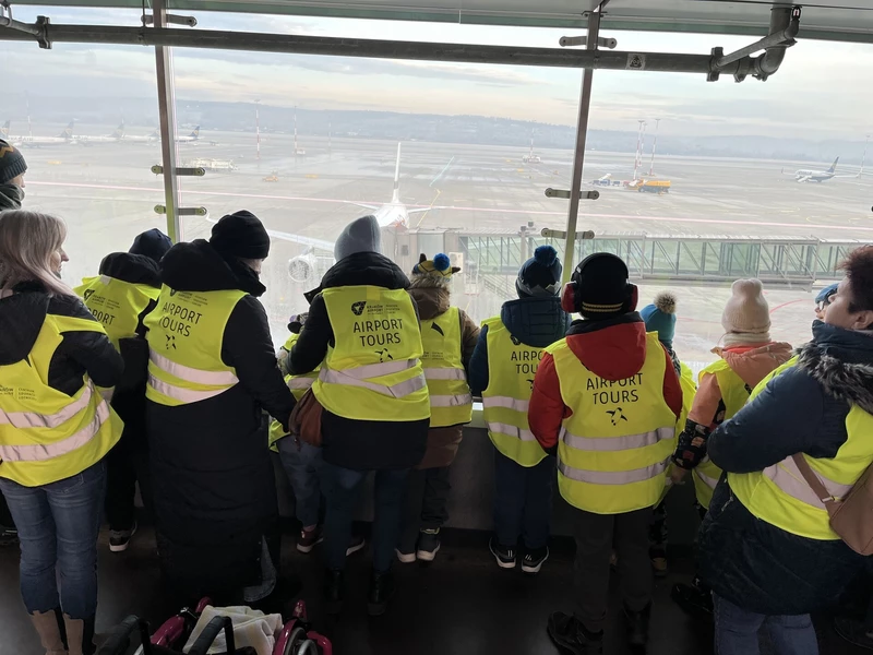 Grupa uczniów w odblaskowych kamizelkach odwrócona tyłem ogląda start samolotu z tarasu widokowego na lotnisku w Balicach.