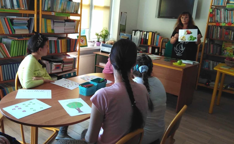 uczennice siedzą przy stoliku w bibliotece, patrzą w stronę bibliotekarza pokazującego ilustracje jabłka w książce.