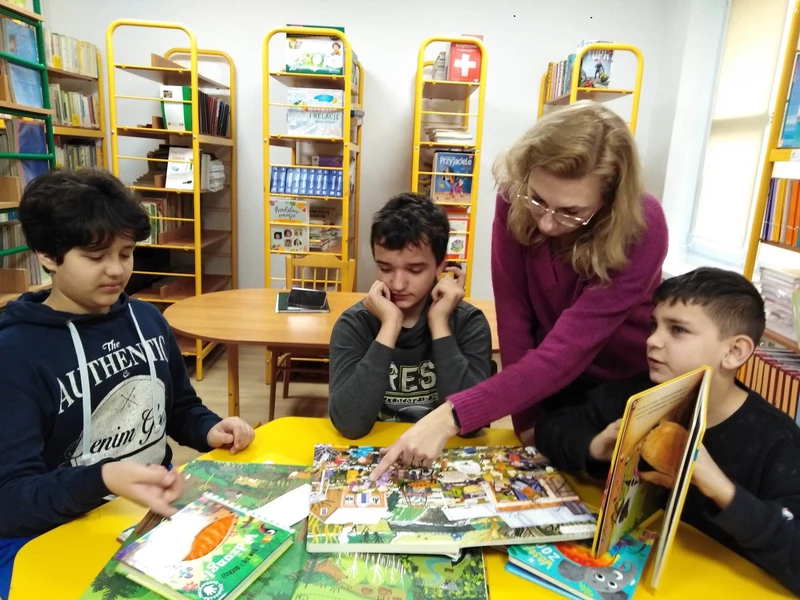 Troje uczniów siedzi przy żółtym stoliku w bibliotece szkolnej. Na stole leżą książki, w których pokazuje coś nauczyciel pochylający się nad uczniami.