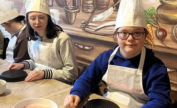 Dwie dziewczyny w czapkach kucharskich i fartuchachsiedzą przy stole, przed nimi leżą okrągłe formy na ciasto.