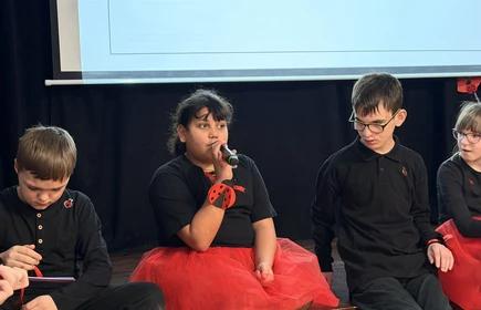 Dziewczynka siedzi na schodach,  mówi do mikrofonu, obok siedzi dwóch chłopców.