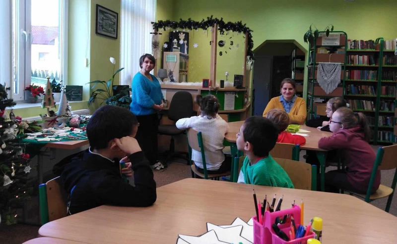 Grupa małych uczniów siedzi przy stolikach w bibliotece szkolnej , patrzą na kobiete która stoi przed nimi i coś opowiada.