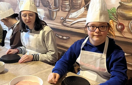 Dwie dziewczyny w czapkach kucharskich i fartuchachsiedzą przy stole, przed nimi leżą okrągłe formy na ciasto.