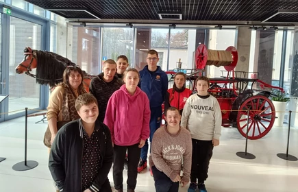 Grupa uczniów stoi w sali wystawowej muzealnej, w tle widać stary pojazd pożarniczy.