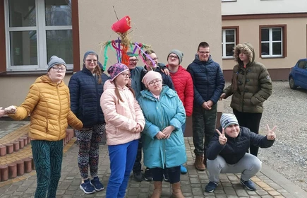 Uczniowie w ciepłej odzieży wierzchniej pozują do zdjęcia na tle budynku szkoły. Jeden uczeń trzyma w ręku kukłę Marzanny.