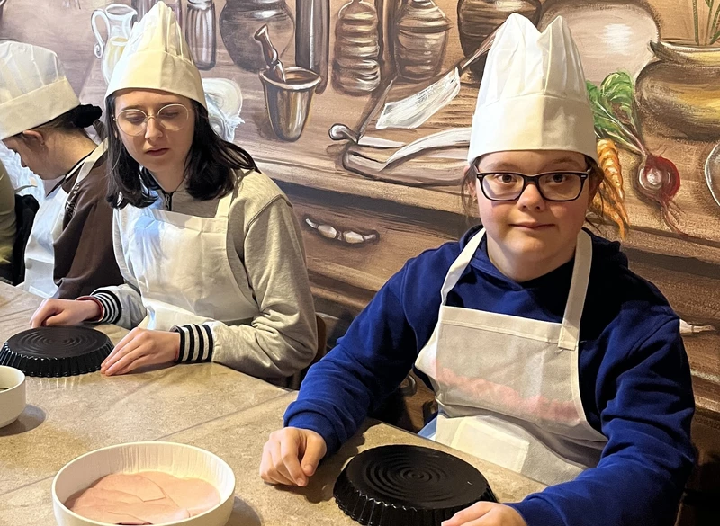 Dwie dziewczyny w czapkach kucharskich i fartuchachsiedzą przy stole, przed nimi leżą okrągłe formy na ciasto.