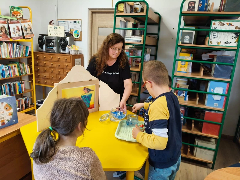 W bibliotece szkolnej nauczyciel pokazuje bajkę japońską dzieciom, które siedzą przy żółtym stoliku.