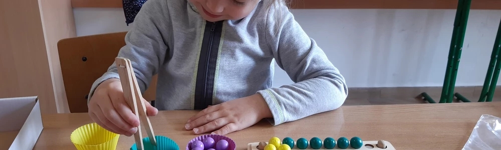 Zdjecie przedstawia dziecko -dziewczynkę , kóra podczas zajęć na lekcji przenosi kolorowe kulki na plansze.