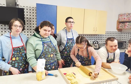 Uczniowie ubrani w fartuchy kuchenne stoją przy blacie, jedna dziewczynka wałkuje ciasto.