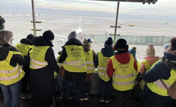 Grupa uczniów w odblaskowych kamizelkach odwrócona tyłem ogląda start samolotu z tarasu widokowego na lotnisku w Balicach.