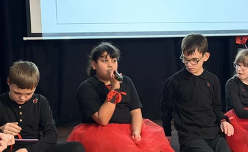 Dziewczynka siedzi na schodach,  mówi do mikrofonu, obok siedzi dwóch chłopców.