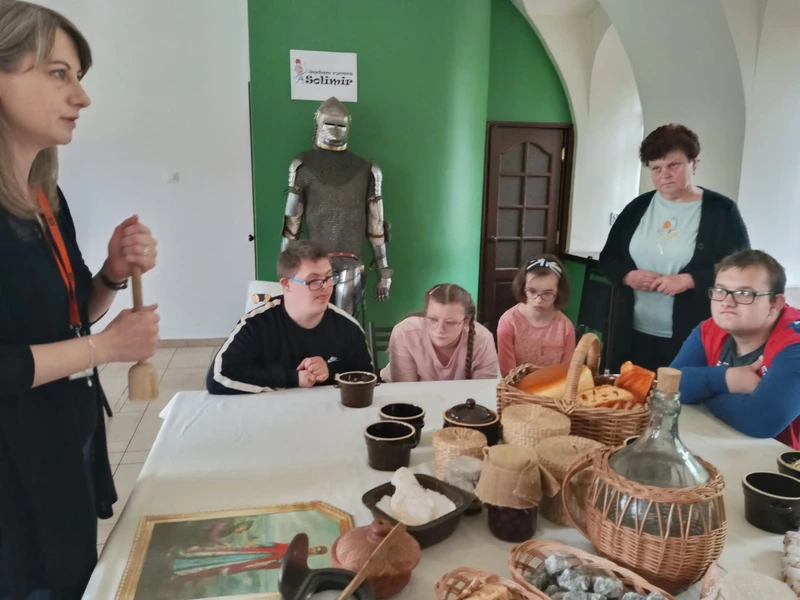 Młodzież siedzi wokół stołu, na którym stoją drewniane kuchenne przedmioty,  gliniane naczynia. Dwie kobiety stoja obok stołu i nachylają się nad nim.