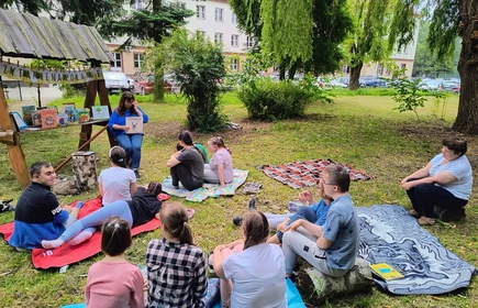 Grupa uczniów siedzi na trawie i słucha treści czytanej książki.