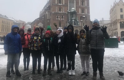 Grupa młodzieży ubranych w  grube czapki i kurtki pozuje do zdjęcia na tle Kościoła Mariackiego na RYnku Głównym w Krakowie, w zimowej scenerii.